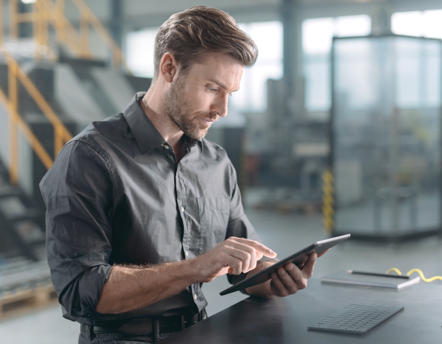 Businessmann steht in einer unscharf zu sehenden Werkshalle und arbeitet am Tablet.
