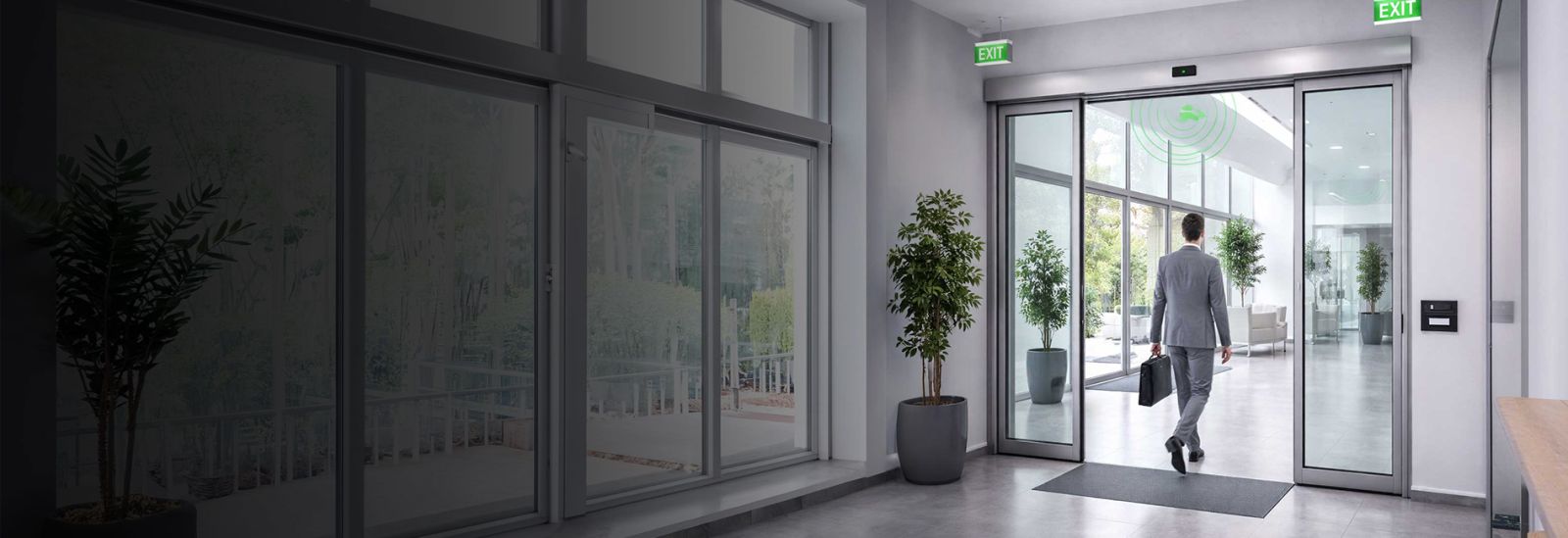Innenbereich eines modernen Gebäudes mit großen Glasfronten, automatischer Schiebetür und mehreren Pflanzen in Töpfen; eine Person geht durch die geöffnete Tür nach draußen.
